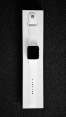 A white smartwatch with a rectangular black screen and a sleek, minimalist design is placed on a white background. The watch band is made of flexible material with multiple adjustment holes for sizing.