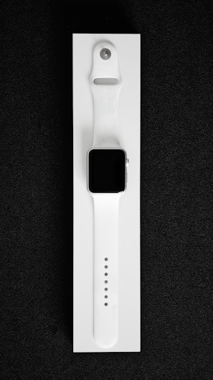 A white smartwatch with a rectangular black screen and a sleek, minimalist design is placed on a white background. The watch band is made of flexible material with multiple adjustment holes for sizing.