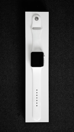 A white smartwatch with a rectangular black screen and a sleek, minimalist design is placed on a white background. The watch band is made of flexible material with multiple adjustment holes for sizing.