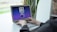 A person is typing on a laptop with a screen displaying a blockchain logo and the word ‘connecting’. The background features a blurred plant and some shelves.
