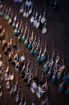 A collection of intricately designed earrings displayed on a dark board. The earrings feature various styles and designs, including spirals, round shapes, and dangling swirls. The colors are a mix of metallic silver and turquoise blue with some white elements, creating an artistic and elegant assortment.