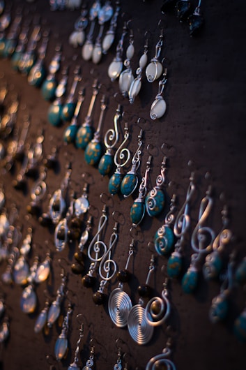 A collection of intricately designed earrings displayed on a dark board. The earrings feature various styles and designs, including spirals, round shapes, and dangling swirls. The colors are a mix of metallic silver and turquoise blue with some white elements, creating an artistic and elegant assortment.