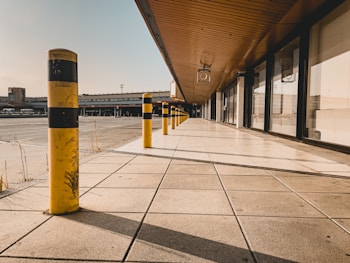 A series of yellow and black striped bollards line the edge of a tiled walkway that runs parallel to a large building with glass windows and a wooden overhang. The surroundings include an open, paved area with infrastructure in the background, under a clear blue sky.