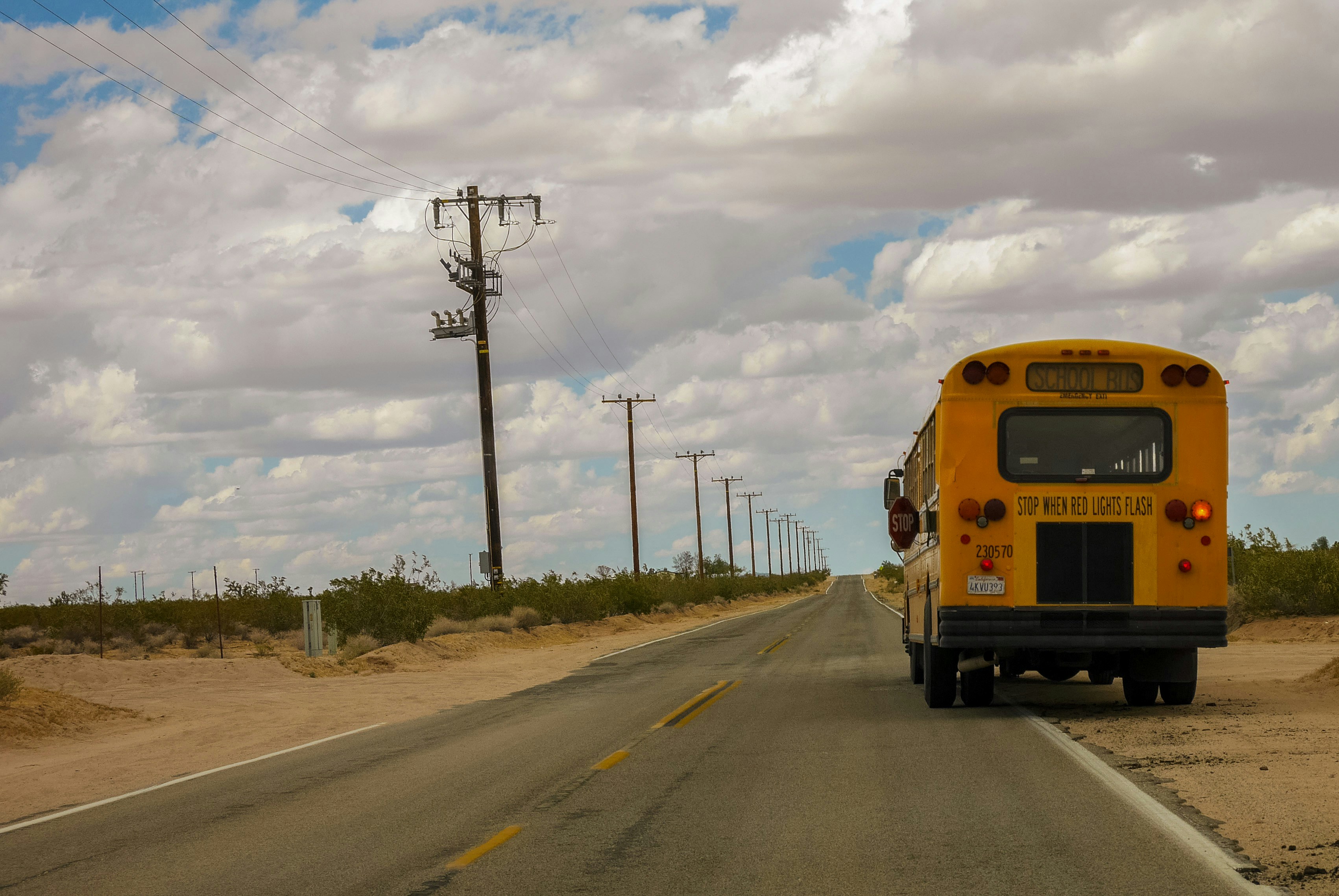 School bus pulled to the side of a two lane road in sandy area. Power poles along other side of road. 