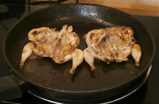Two small birds, likely quail, are cooking in a dark, round frying pan. The birds have been split open and are being seared, showing a grilled texture with some browning on the skin.