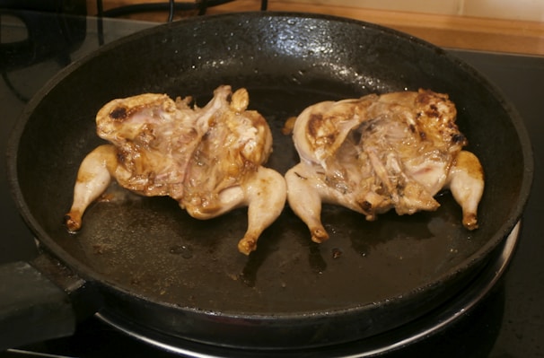 Two small birds, likely quail, are cooking in a dark, round frying pan. The birds have been split open and are being seared, showing a grilled texture with some browning on the skin.