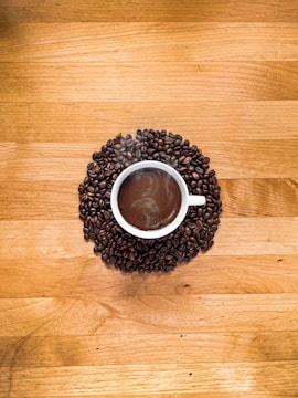 A white cup filled with steaming hot coffee is surrounded by a circle of dark roasted coffee beans, placed on a wooden table with a warm, natural finish.