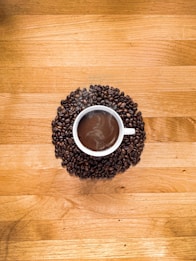A white cup filled with steaming hot coffee is surrounded by a circle of dark roasted coffee beans, placed on a wooden table with a warm, natural finish.