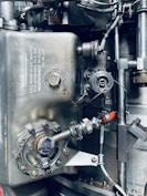 A detailed view of an industrial mechanical system, featuring metallic components such as pipes, valves, and bolts arranged in a complex structure. The surface of the metal parts is marked with technical inscriptions and a few spots of rust. Wires and connectors run alongside the metal parts, suggesting functionality related to machinery or engineering.