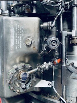 A detailed view of an industrial mechanical system, featuring metallic components such as pipes, valves, and bolts arranged in a complex structure. The surface of the metal parts is marked with technical inscriptions and a few spots of rust. Wires and connectors run alongside the metal parts, suggesting functionality related to machinery or engineering.