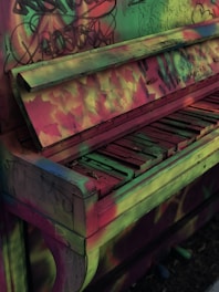 A vintage upright piano with colorful graffiti and street art covering its surface. The keys seem worn and some are missing, while the entire piano exhibits a blend of vibrant green, pink, and yellow hues.