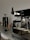 A sleek coffee machine with a polished silver and black finish sits prominently on a countertop. In the background, various coffee-making accessories, including a grinder and a pour-over kettle, are arranged neatly, suggesting a setup for making specialty coffee.