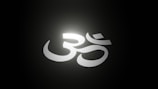 A white Om symbol is illuminated against a dark background, creating a glowing effect with a soft halo of light around it.