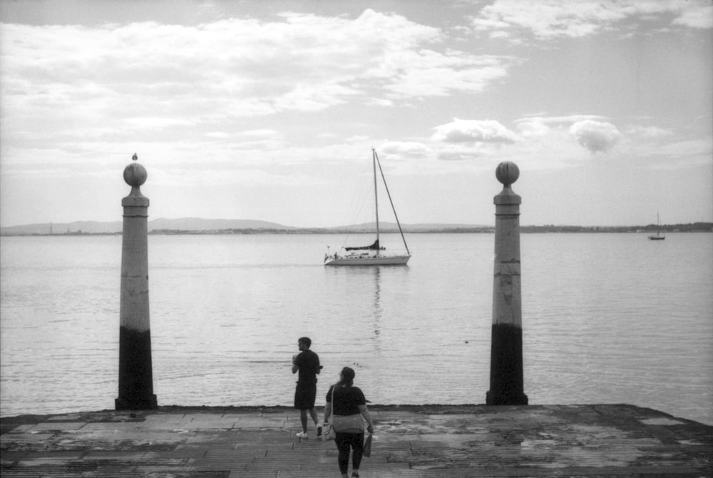 a black and white photo of two people walking on a pier