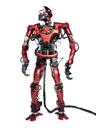 A humanoid robot with a sleek red metallic body and exposed wiring stands on a white background. It has a complex mechanical structure with various components and joints visible, including multiple wiring and cables. The robot's head includes a visor-like screen, and it appears to be equipped with various sensors and tools.
