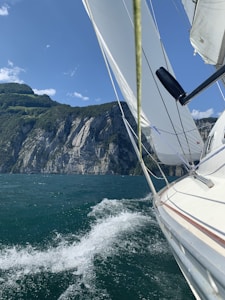 A sailboat is navigating through clear, blue waters with its sails fully deployed. The boat is leaning to the side, creating waves and a dynamic splash. In the background, there are steep, rocky cliffs covered with patches of green vegetation under a bright blue sky with scattered clouds.