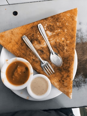 A large triangular dosa is placed on a white plate, accompanied by two small bowls of sauces, one orange and one cream-colored. A fork and spoon are resting on top of the dosa.
