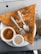 A large triangular dosa is placed on a white plate, accompanied by two small bowls of sauces, one orange and one cream-colored. A fork and spoon are resting on top of the dosa.