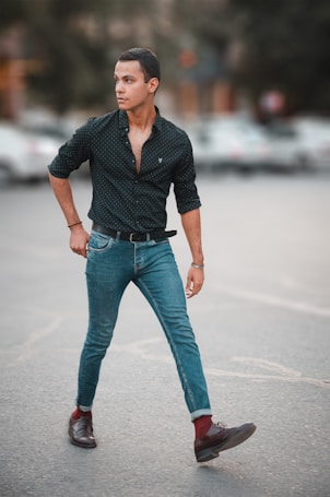A person walks confidently across a street, dressed in a black patterned shirt and slim-fit jeans. The background is blurred, highlighting the individual. The person appears focused, looking to the side with hands casually placed in pockets.