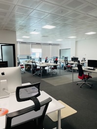 A modern office space with multiple desks and chairs, each equipped with computers and office supplies. The room is well-lit with natural light coming in through large windows with blinds. The ceiling has a grid pattern with recessed lights, and the space appears organized and tidy.