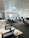 A modern office space with multiple desks and chairs, each equipped with computers and office supplies. The room is well-lit with natural light coming in through large windows with blinds. The ceiling has a grid pattern with recessed lights, and the space appears organized and tidy.