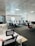 A modern office space with multiple desks and chairs, each equipped with computers and office supplies. The room is well-lit with natural light coming in through large windows with blinds. The ceiling has a grid pattern with recessed lights, and the space appears organized and tidy.
