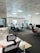 A modern office space with multiple desks and chairs, each equipped with computers and office supplies. The room is well-lit with natural light coming in through large windows with blinds. The ceiling has a grid pattern with recessed lights, and the space appears organized and tidy.