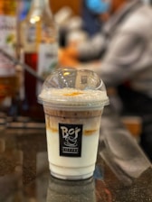 A plastic cup filled with an iced coffee beverage featuring layers of milk and coffee, topped with a clear plastic dome lid. The cup bears a logo reading 'Bo's Coffee'. The background is blurred, showing indistinct figures and bottles.