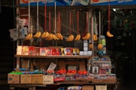 A small market stall displays a variety of items, including bunches of bananas hanging from strings. There are colorful packages featuring images of people stacked on the shelf, alongside boxes of bottled drinks and fresh tomatoes. The scene includes other household or small retail items.