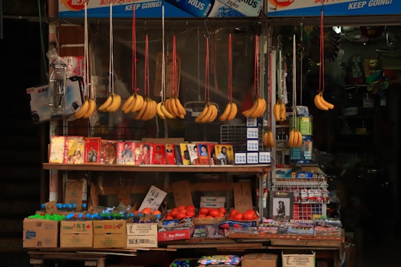 A small market stall displays a variety of items, including bunches of bananas hanging from strings. There are colorful packages featuring images of people stacked on the shelf, alongside boxes of bottled drinks and fresh tomatoes. The scene includes other household or small retail items.