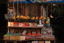 A small market stall displays a variety of items, including bunches of bananas hanging from strings. There are colorful packages featuring images of people stacked on the shelf, alongside boxes of bottled drinks and fresh tomatoes. The scene includes other household or small retail items.