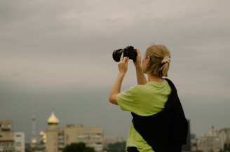 A person with blonde hair stands on a rooftop holding a camera and taking a photograph. The person is wearing a light green shirt with a black jacket or cloth draped over their shoulders. In the background, there are several buildings, one with a distinctive gold dome, and the sky is overcast with shades of gray.