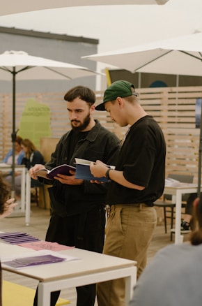 Two individuals are standing at a table, intently looking at and discussing what appears to be purple brochures or booklets. One person is wearing black clothing and the other has a green cap. They are in an open space with tables, chairs, and large white umbrellas above. There is a wooden fence in the background.