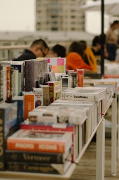 A table filled with various books arranged closely together in an outdoor setting, with several people in the background engaged with the items on the table. The books are diverse in subject, with prominent titles visible. The scene is set on a rooftop or a terrace, with a blurred cityscape in the background.