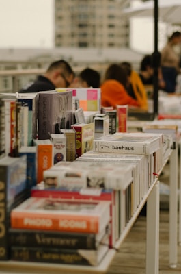 A table filled with various books arranged closely together in an outdoor setting, with several people in the background engaged with the items on the table. The books are diverse in subject, with prominent titles visible. The scene is set on a rooftop or a terrace, with a blurred cityscape in the background.