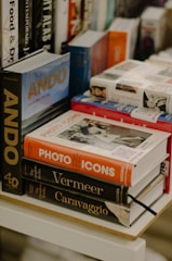 An assortment of books arranged on a wooden table or shelf. The titles are diverse and include 'ANDO', 'Photo Icons', 'Vermeer', and 'Caravaggio'. The books vary in size and color, with some stacked neatly and others leaning against each other. The background contains additional books, possibly indicating a bookstore or library setting.