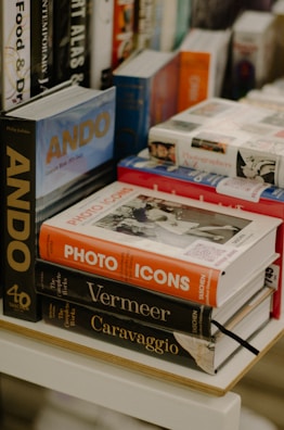 An assortment of books arranged on a wooden table or shelf. The titles are diverse and include 'ANDO', 'Photo Icons', 'Vermeer', and 'Caravaggio'. The books vary in size and color, with some stacked neatly and others leaning against each other. The background contains additional books, possibly indicating a bookstore or library setting.