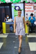 A person confidently walks down a runway in a striped dress and black heels. Behind them, there are banners and signs indicating a beauty college event. Other people are present, including a seated person in a mask at a desk and another person in a hat, possibly taking a photo. The setting is decorated with green and white balloons.