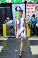 A person confidently walks down a runway in a striped dress and black heels. Behind them, there are banners and signs indicating a beauty college event. Other people are present, including a seated person in a mask at a desk and another person in a hat, possibly taking a photo. The setting is decorated with green and white balloons.