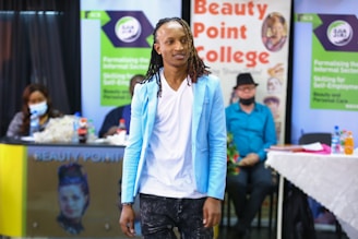 A person with long braided hair is wearing a light blue blazer over a white shirt and dark pants. They are standing in front of a background that includes branding for Beauty Point College. There is a seated audience and tables with bottled drinks in the background.