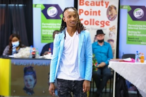 A person with long braided hair is wearing a light blue blazer over a white shirt and dark pants. They are standing in front of a background that includes branding for Beauty Point College. There is a seated audience and tables with bottled drinks in the background.