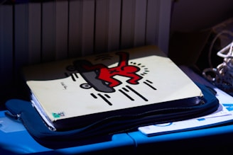A folder with a distinctive graphic artwork featuring a red figure on a skateboard against a cream background. The folder rests atop a blue surface, with other materials partially visible underneath. The scene is contrasted by shadows.