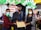 Three individuals are wearing masks. The person in the center is dressed in a graduation cap and gown, holding a certificate. The setting includes decorations with balloons and a banner in the background indicating it might be a graduation event.