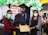 Three individuals are wearing masks. The person in the center is dressed in a graduation cap and gown, holding a certificate. The setting includes decorations with balloons and a banner in the background indicating it might be a graduation event.