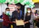 Three individuals are wearing masks. The person in the center is dressed in a graduation cap and gown, holding a certificate. The setting includes decorations with balloons and a banner in the background indicating it might be a graduation event.