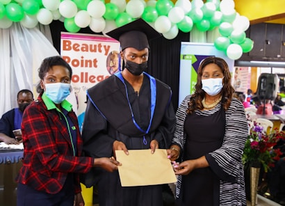 Three individuals are wearing masks. The person in the center is dressed in a graduation cap and gown, holding a certificate. The setting includes decorations with balloons and a banner in the background indicating it might be a graduation event.