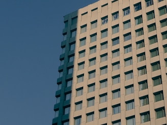 A modern high-rise building with a facade made of concrete panels and numerous rectangular windows, set against a clear blue sky. Japanese characters are visible at the top of the building.