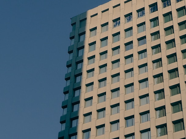 A modern high-rise building with a facade made of concrete panels and numerous rectangular windows, set against a clear blue sky. Japanese characters are visible at the top of the building.