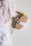 A wooden brush with a white fabric strap lies next to a sprig of small yellow flowers, all placed on a soft, light-colored surface. Draped fabric, in shades of soft pink and white, gently fall on the left side of the image.