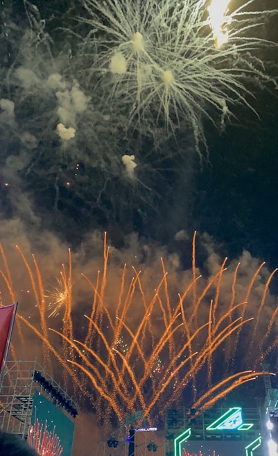 Bright fireworks burst with orange and white sparks against a dark night sky, illuminating a large crowd below. Various patterns of streaks and explosions create a festive and vibrant scene. The lower part of the image shows structures and a digital screen displaying lights and graphics.